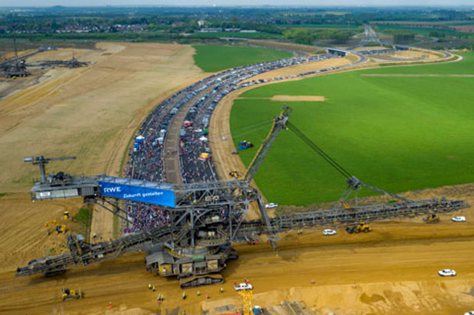 Eine Luftaufnahme eines massiven Baggers mit einem langen Förderband in einem Feld, umgeben von Protestierenden und Fahrzeugen auf einer Straße.