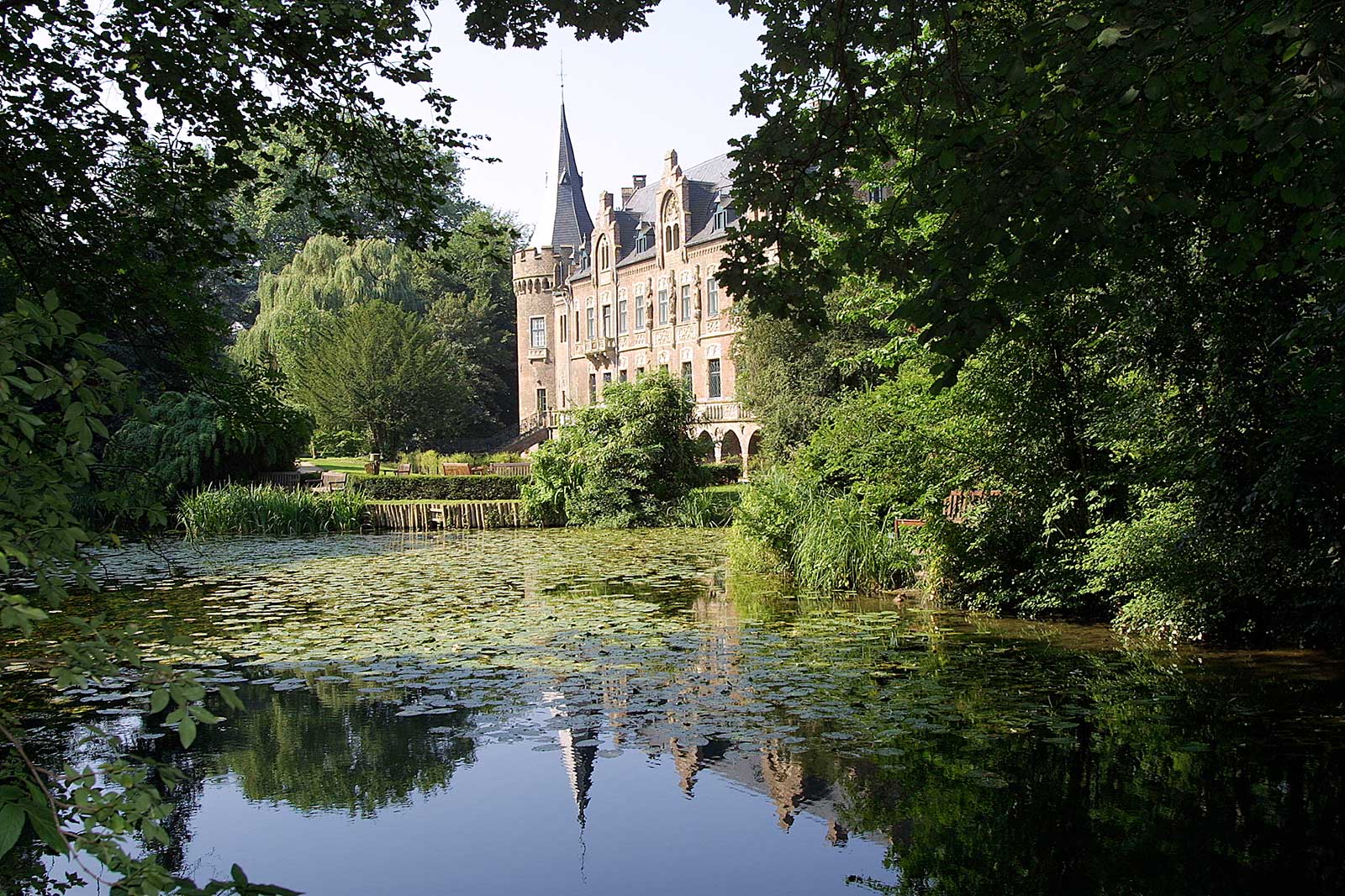 Ein ruhiger Seerosenteich im Vordergrund spiegelt ein helles, steinernes Herrenhaus mit Türmchen, umgeben von dichtem, grünem Laub unter blauem Himmel.