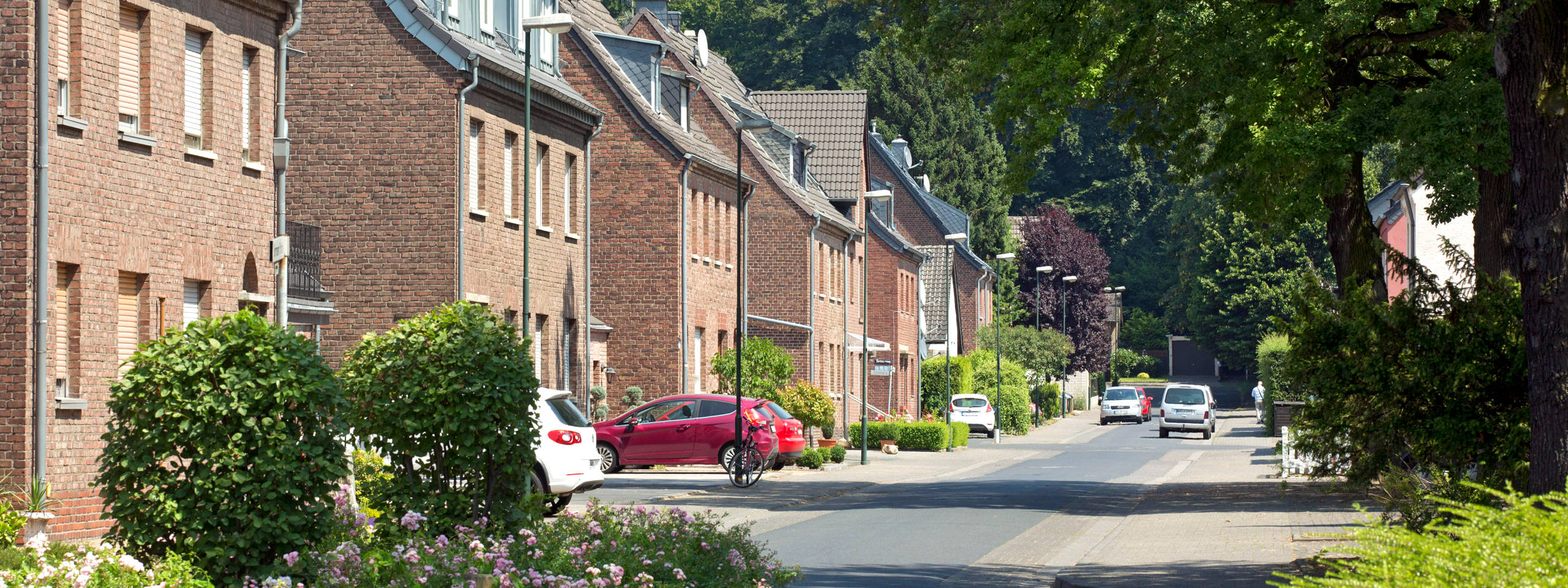 Eine ruhige Straße mit Backsteinhäusern, grünen Bäumen und Fahrzeugen, die entlang der Straße parken.
