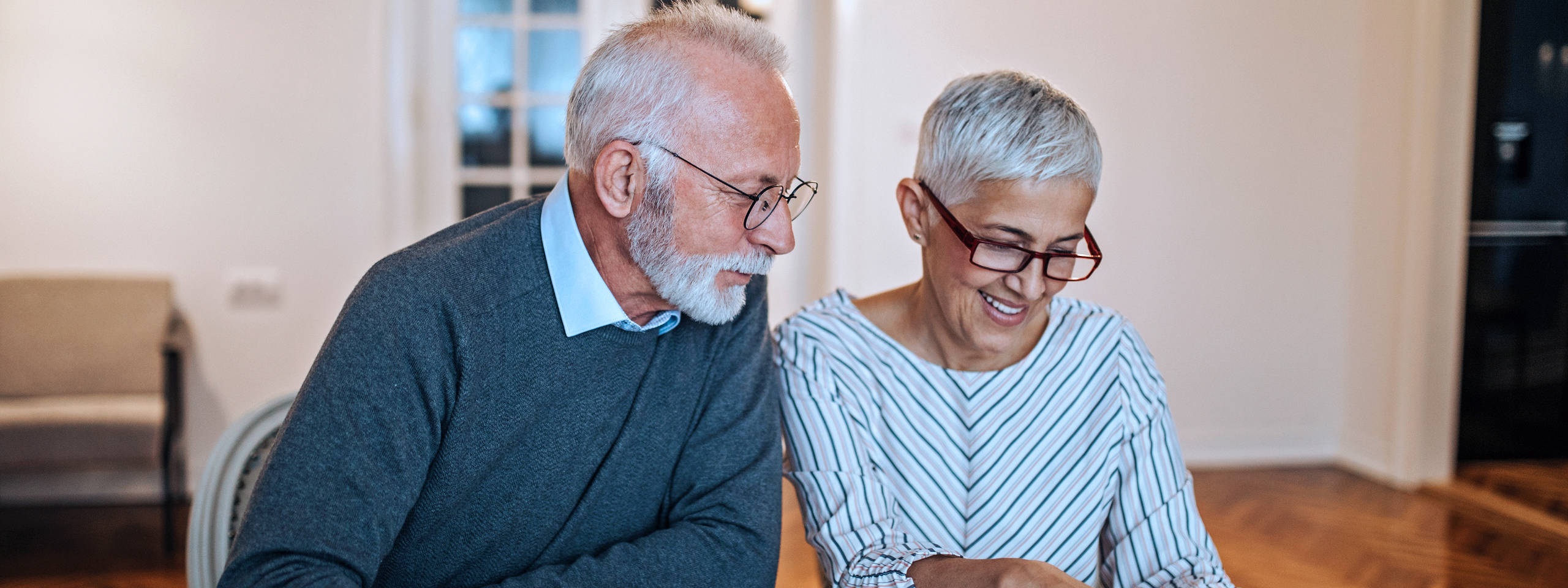 Ein älteres Paar sitzt zusammen an einem Tisch, lächelnd und sich unterhaltend, in einem hellen, modernen Raum.