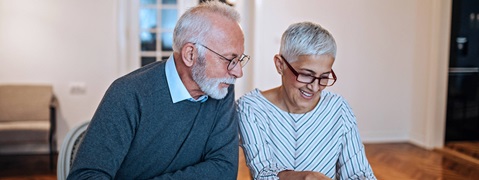 Ein älteres Paar sitzt zusammen an einem Tisch, lächelnd und sich unterhaltend, in einem hellen, modernen Raum.