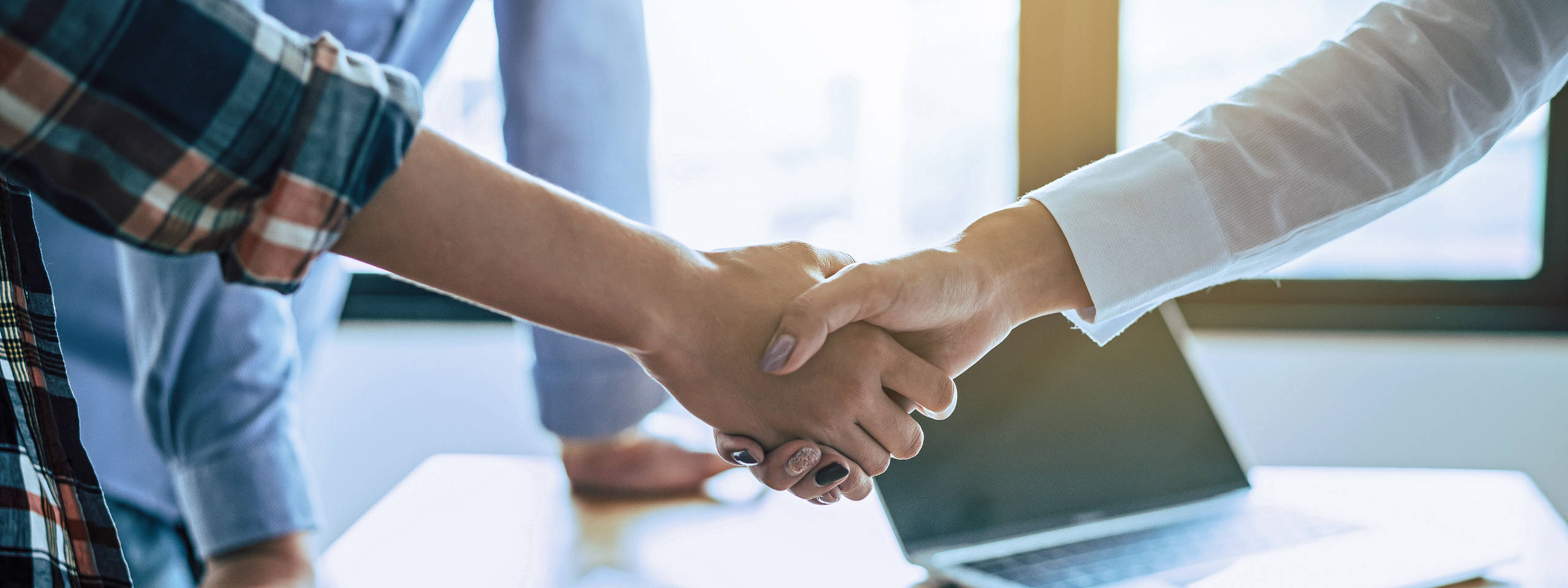 Zwei Personen schütteln sich die Hände in einem modernen Büro mit einem Laptop im Hintergrund.