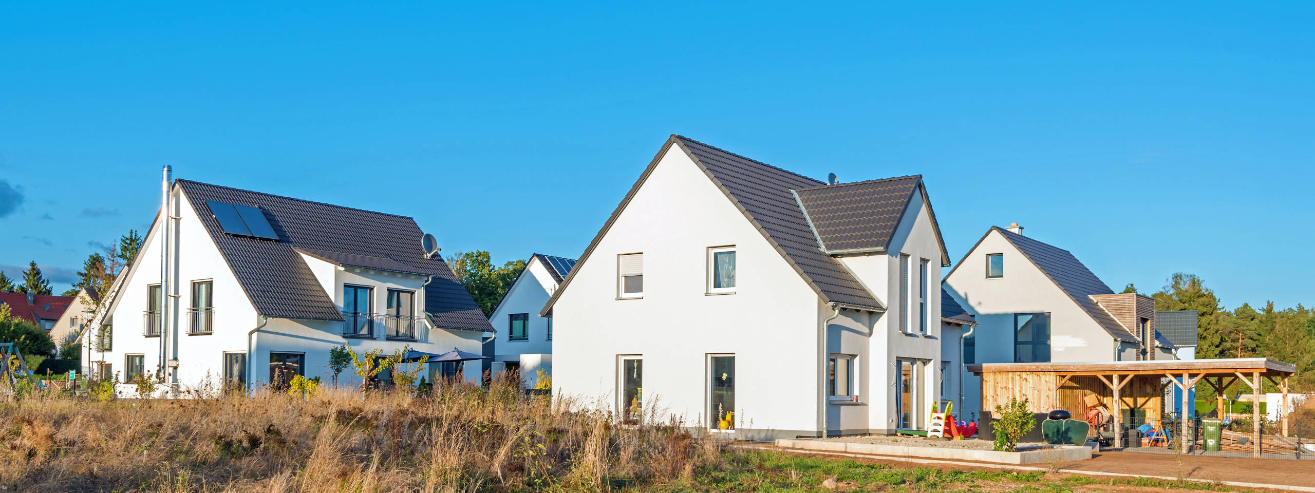 Moderne, weiße Häuser mit dunklen Dächern stehen in einer ruhigen Nachbarschaft. Grünflächen umgeben die Gebäude.