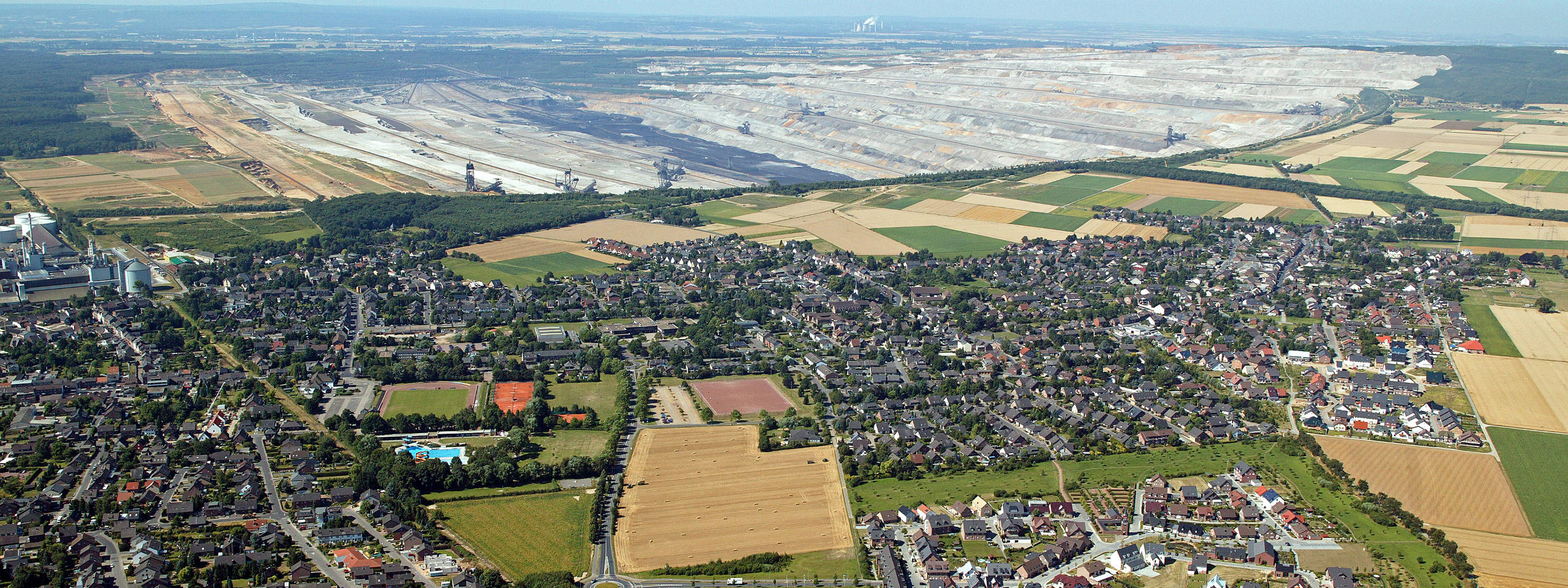 Luftaufnahme eines Dorfes neben einem großen Tagebau und landwirtschaftlich genutzten Feldern.