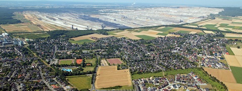 Luftaufnahme eines Dorfes neben einem großen Tagebau und landwirtschaftlich genutzten Feldern.
