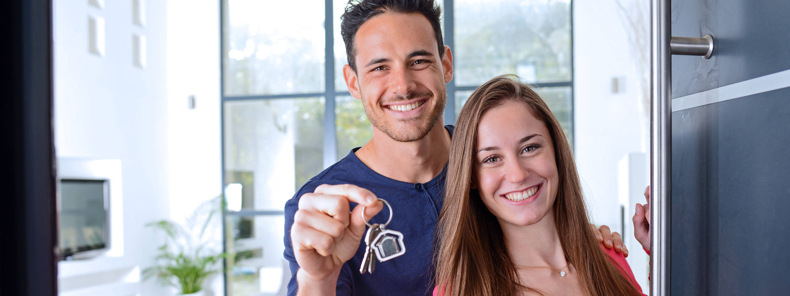 Ein Paar steht an einer Tür und hält einen Schlüsselbund. Der Raum hat große Fenster und moderne Einrichtung.