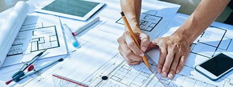 Hände, die einen Plan mit einem Bleistift zeichnen, umgeben von Plänen, einem Tablet und einem Smartphone.