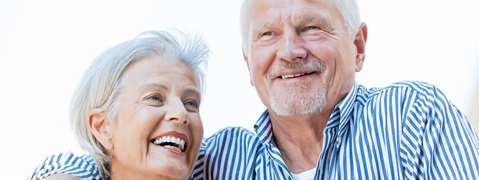 Ein älteres Paar lächelt, trägt blau-weiß gestreifte Hemden und hat einen hellen Hintergrund.