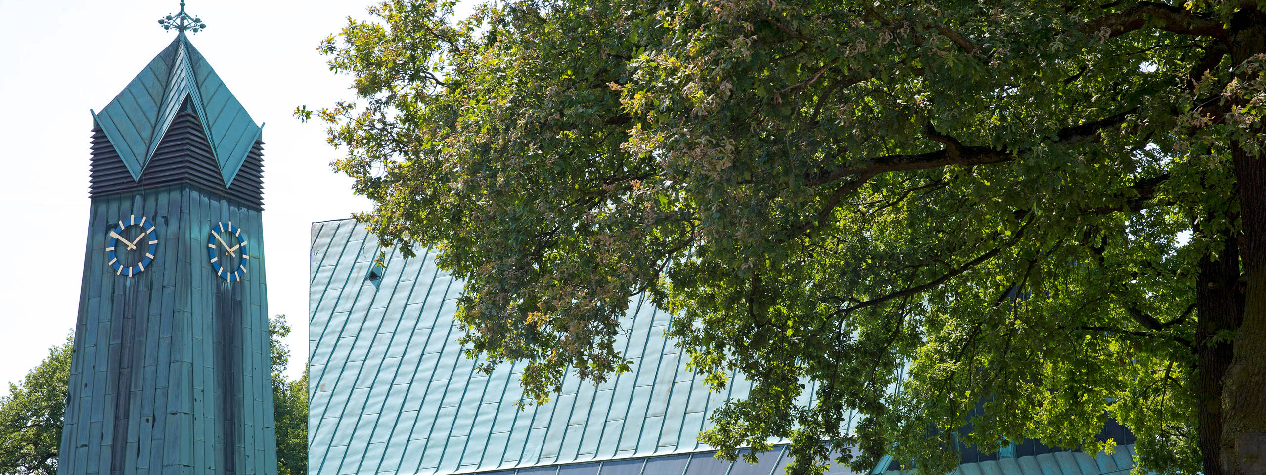 Ein Glockenturm mit bläulich-grüner Fassade und zwei Uhren, umgeben von üppigem Grün und einem modernen Gebäude.