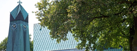 Ein Glockenturm mit bläulich-grüner Fassade und zwei Uhren, umgeben von üppigem Grün und einem modernen Gebäude.