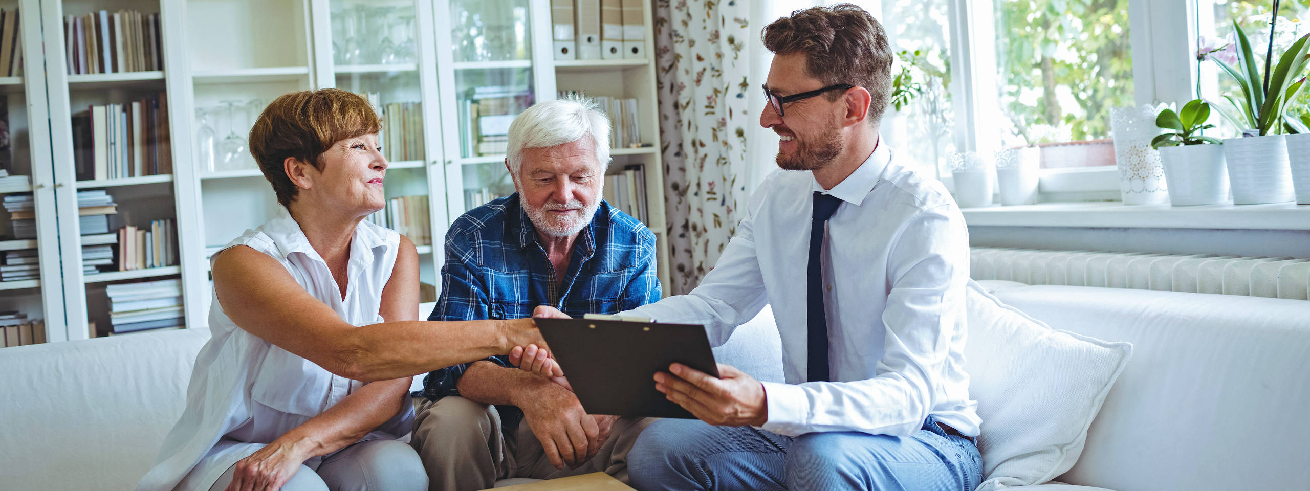 Eine Frau gibt einem Mann die Hand, während ein älterer Mann daneben sitzt. Im Hintergrund sind Bücherregale sichtbar.