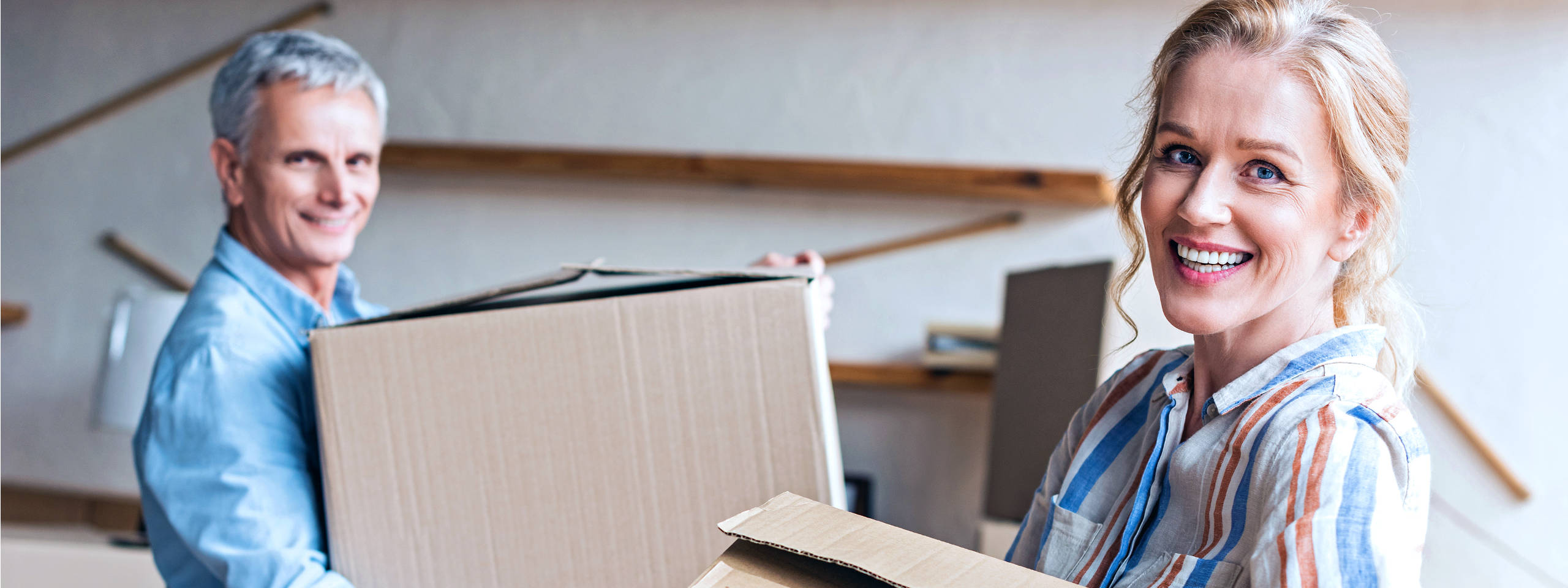 Zwei Personen tragen Kartons in einem modernen Raum mit Holzregalen im Hintergrund.