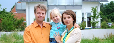Ein lächelndes Elternpaar hält ein Kind in einem blauen T-Shirt. Im Hintergrund steht ein modernes Haus.