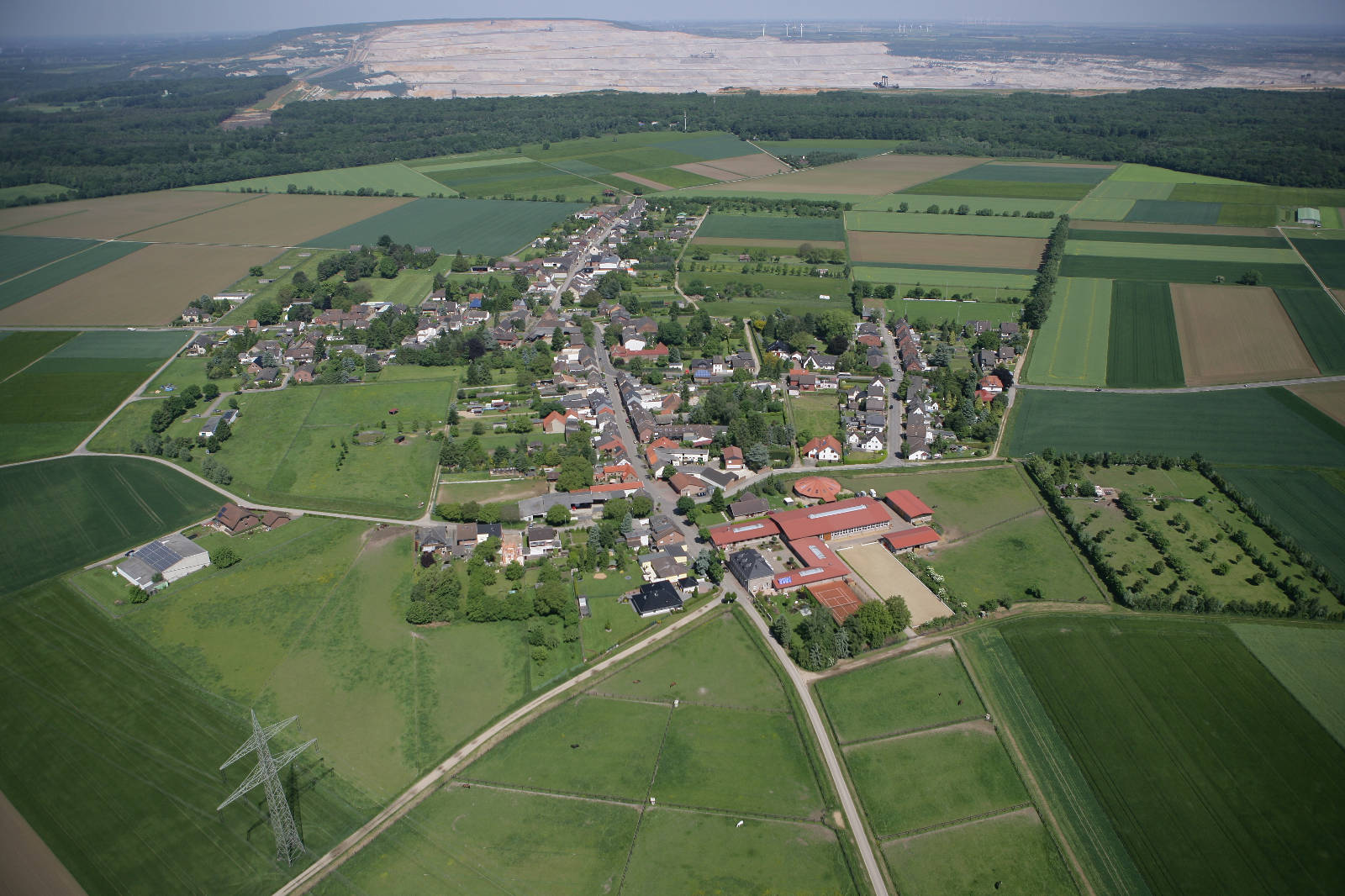 Eine Luftaufnahme eines kleinen Dorfes umgeben von Feldern und Wäldern, im Hintergrund ist ein Tagebau zu sehen.