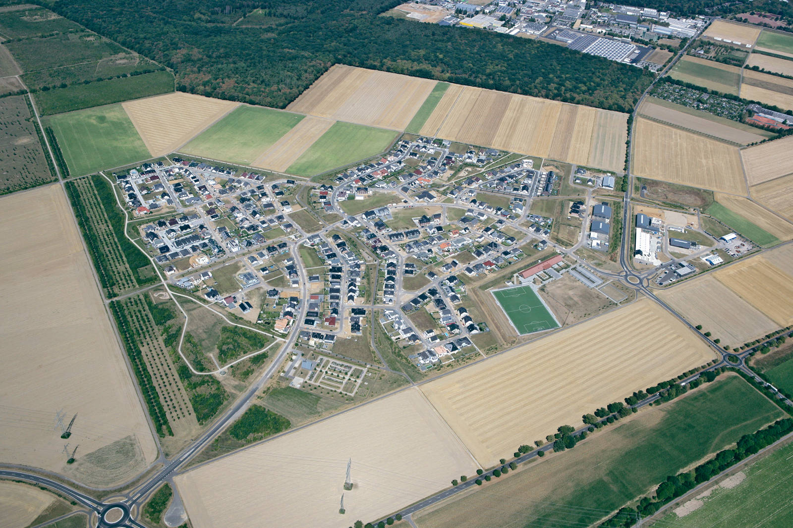 Luftaufnahme einer Wohnsiedlung mit Straßen, Häusern und landwirtschaftlichen Flächen umgeben von Wäldern.