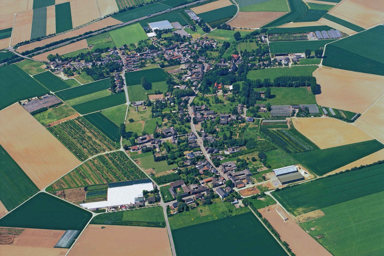 Luftaufnahme eines kleinen Dorfes umgeben von grünen Feldern und landwirtschaftlichen Flächen.