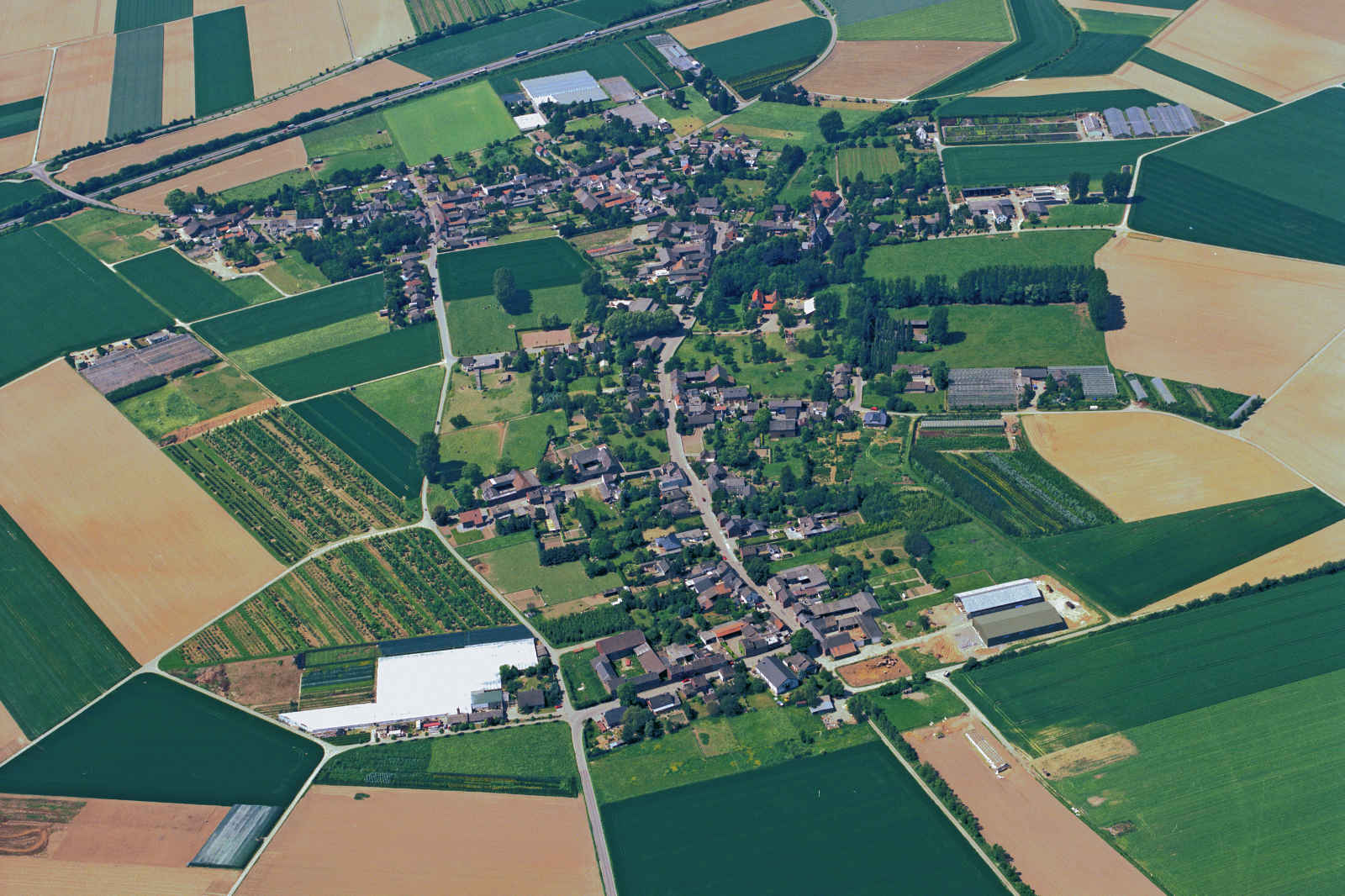 Luftaufnahme eines kleinen Dorfes umgeben von grünen Feldern und landwirtschaftlichen Flächen.