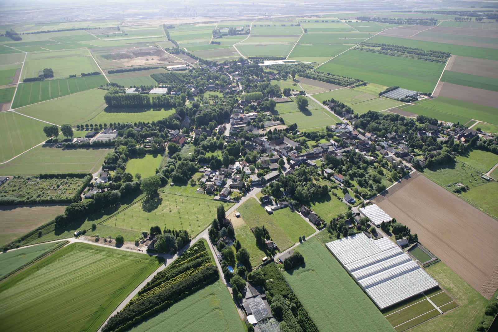 Luftaufnahme eines Dorfes umgeben von grünen Feldern und Ackerland, mit Wohnhäusern und Gewächshäusern sichtbar.