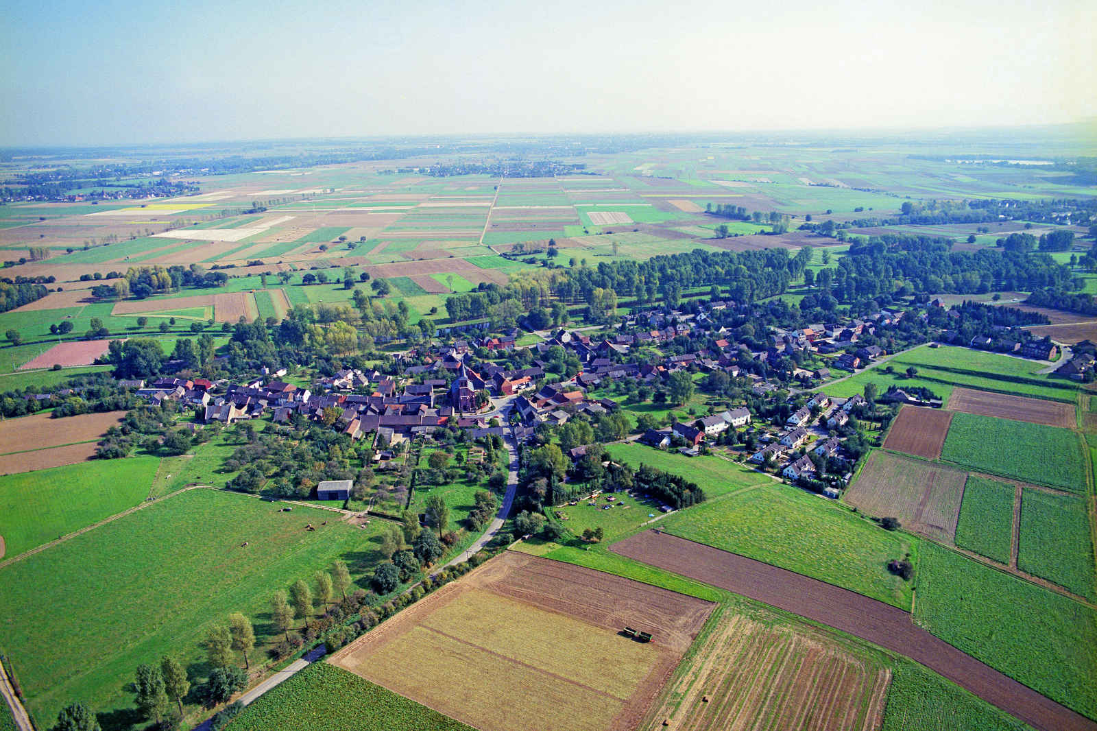 Luftaufnahme eines kleinen Dorfes umgeben von weiten grünen Feldern und Ackerland.