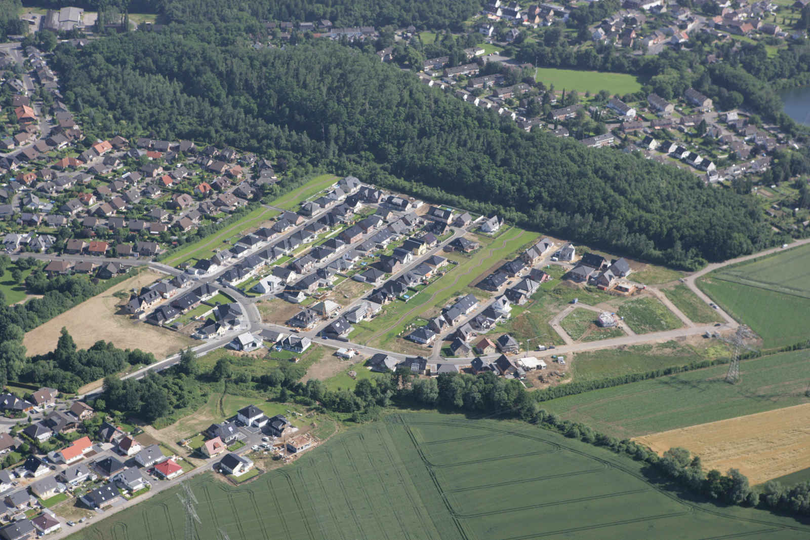 Luftaufnahme eines Neubaugebiets mit unvollständigen Häusern, umgeben von Wäldern und Feldern.