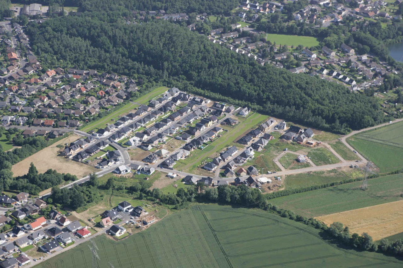 Luftaufnahme eines Neubaugebiets mit unvollständigen Häusern, umgeben von Wäldern und Feldern.