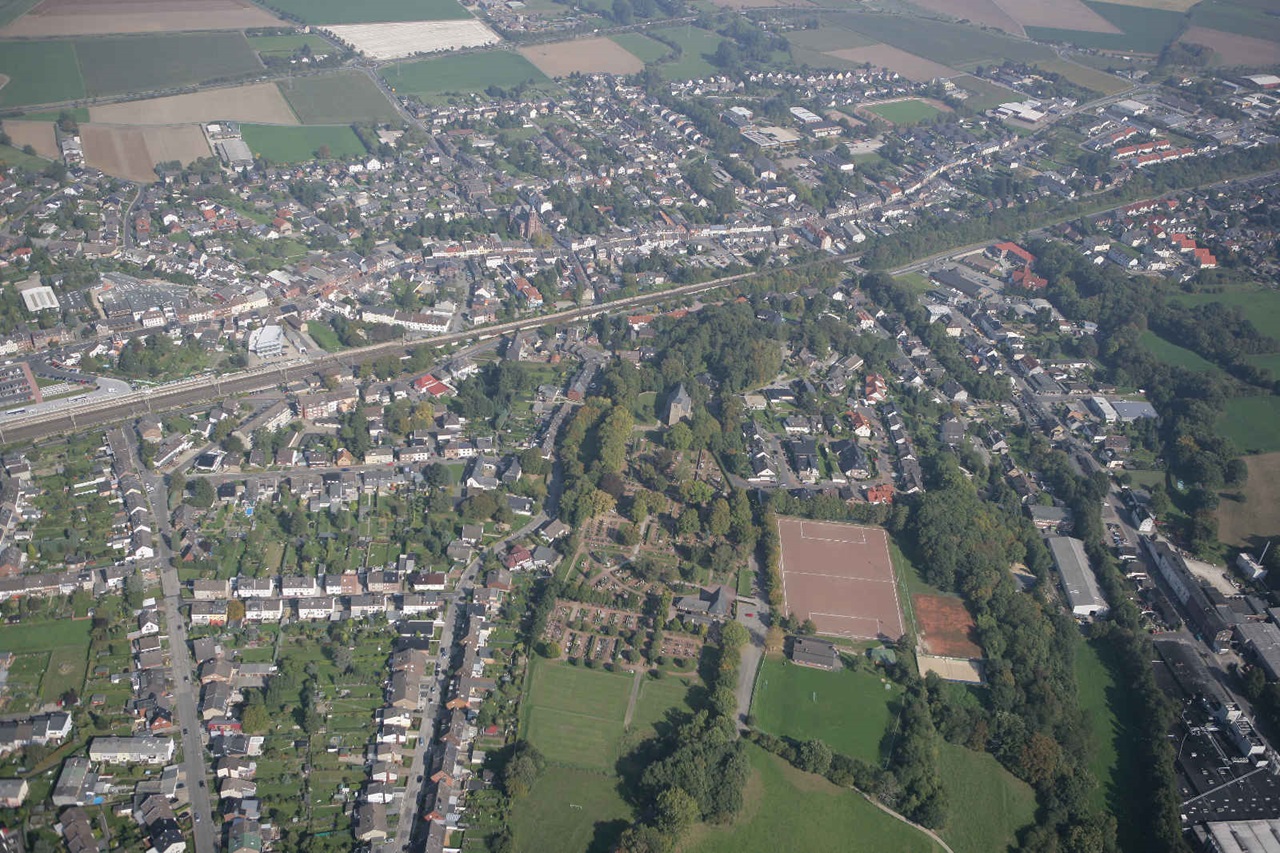 Luftaufnahme einer Stadt mit Wohnhäusern, Straßen, Parks und Feldern. Grüne Flächen und Sportplätze sind sichtbar.
