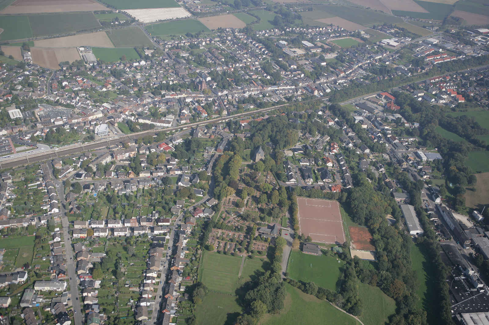 Luftaufnahme einer Stadt mit Wohnhäusern, Straßen, Parks und Feldern. Grüne Flächen und Sportplätze sind sichtbar.