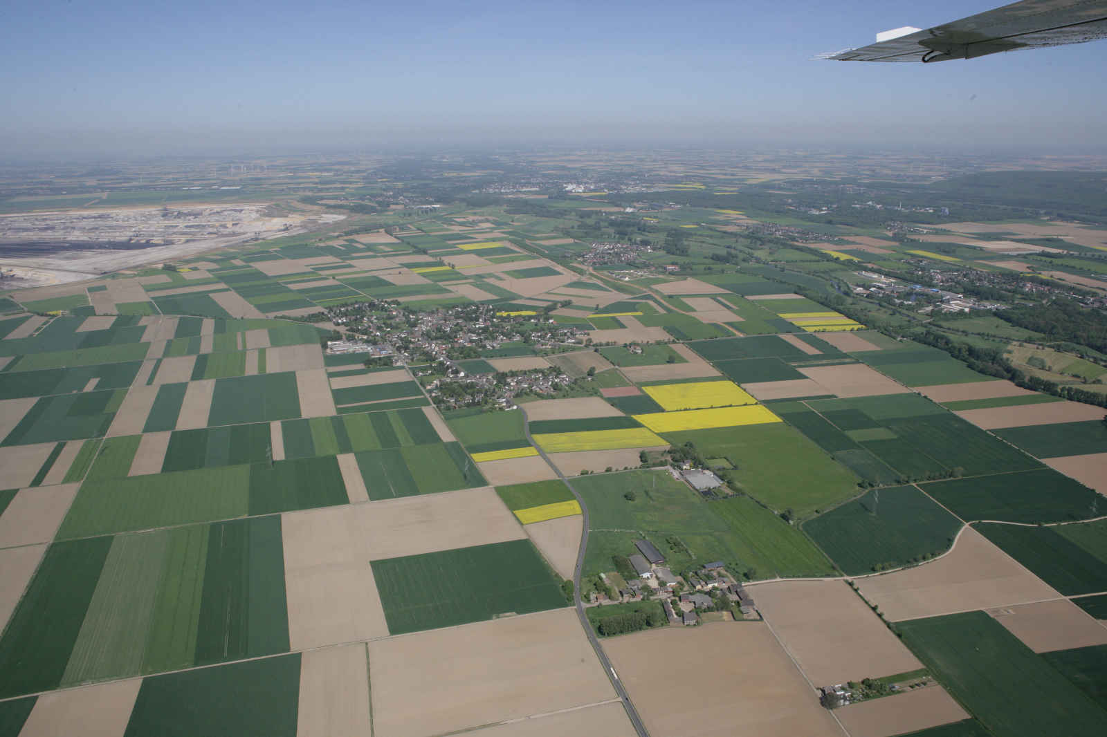 Luftaufnahme eines Flickenteppichs aus grünen und braunen Feldern, mit einem kleinen Dorf inmitten landwirtschaftlicher Flächen.