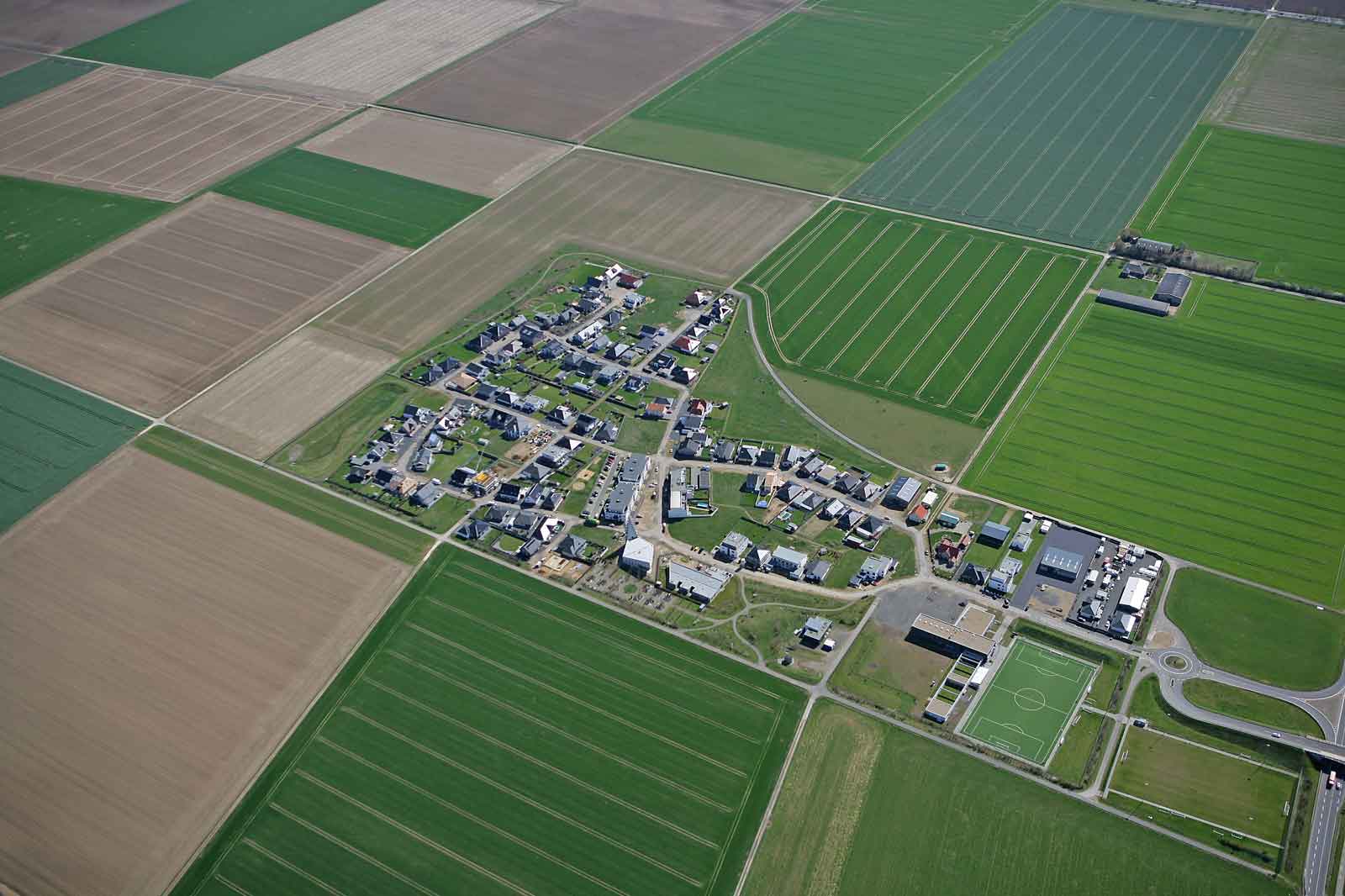 Eine Vogelperspektive auf ein kleines Dorf, umgeben von weiten grünen und braunen Feldern, mit einem Sportplatz sichtbar.