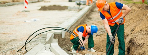 Zwei Bauarbeiter in Sicherheitskleidung graben auf einer Baustelle, im Hintergrund ist Maschinen auszumachen.