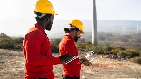 Two workers in hard hats and red shirts stand outdoors, using a tablet and a drone controller, with wind turbines in the background.
