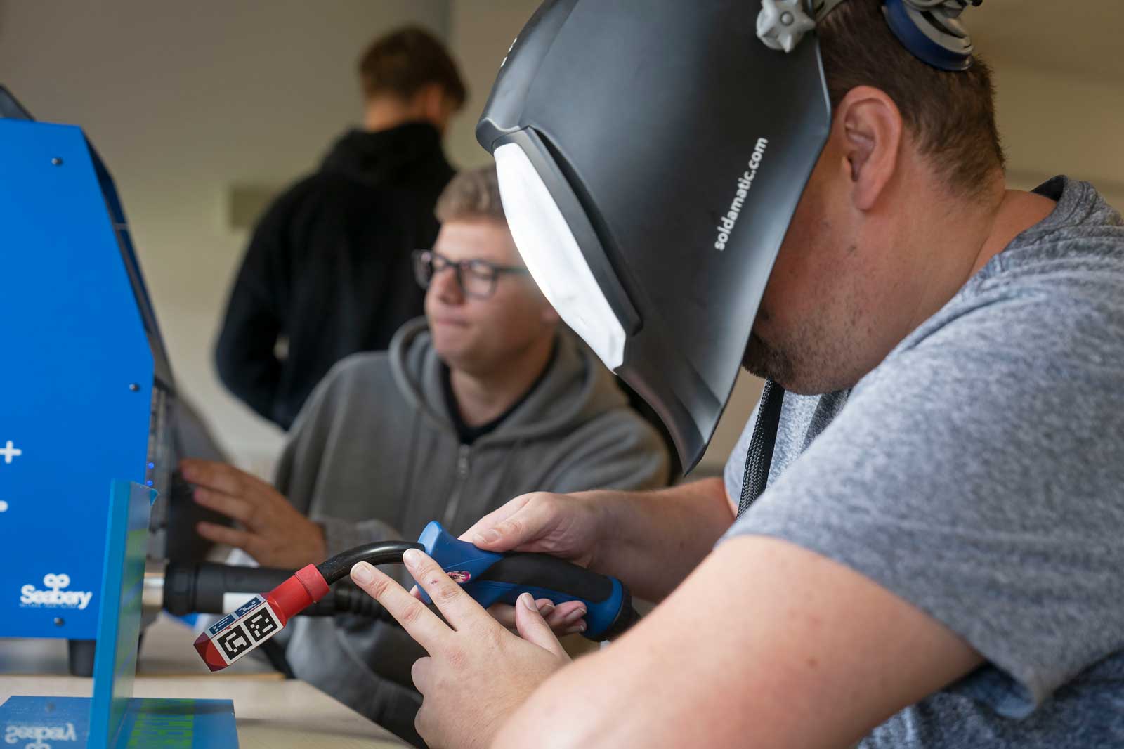 A man is wearing a welding helmet and holding a welding torch. Other people are visible in the background.