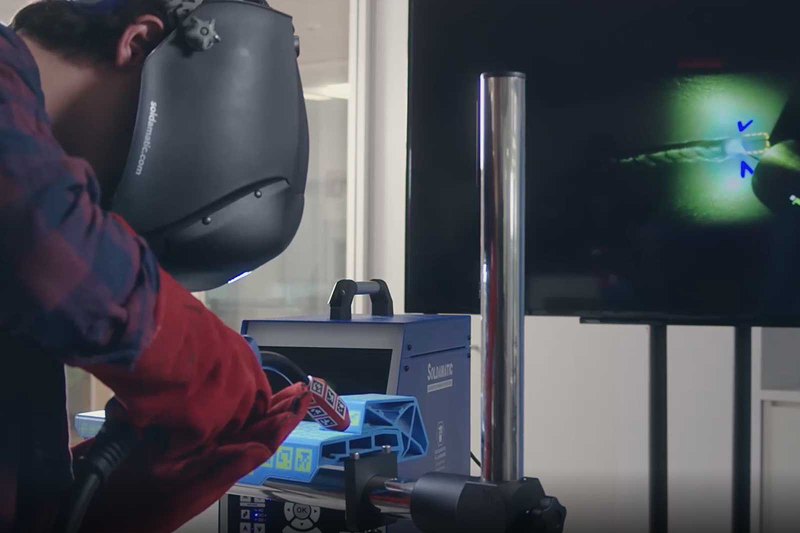 A man in a welding helmet and red gloves is working on a welding machine, while a screen displays the process.