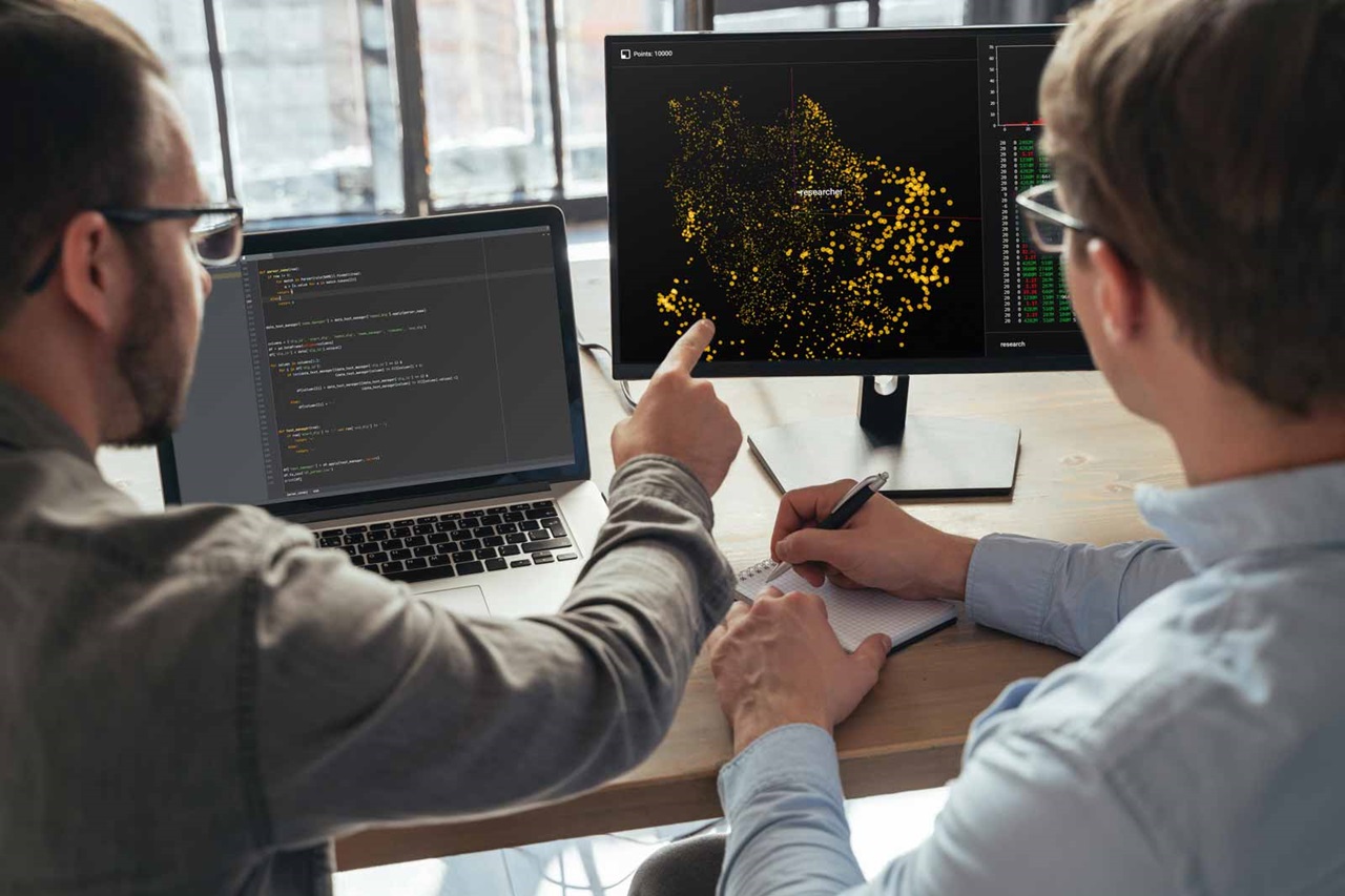 Zwei Personen, die an einem Schreibtisch arbeiten, auf Bildschirme mit Datenvisualisierungen und Programmierer-Code schauen.