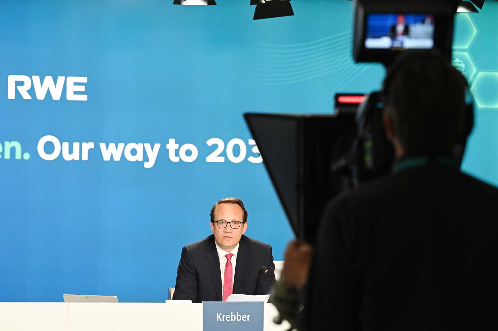 Eine Konferenzszene mit Markus Krebber am Tisch, während ein Kameramann die Präsentation aufnimmt. RWE-Branding ist sichtbar.