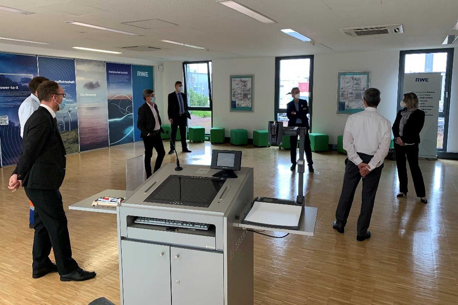 Eine Gruppe von Fachleuten in formeller Kleidung diskutiert in einem hellen Büroraum mit grünen Sitzgelegenheiten.