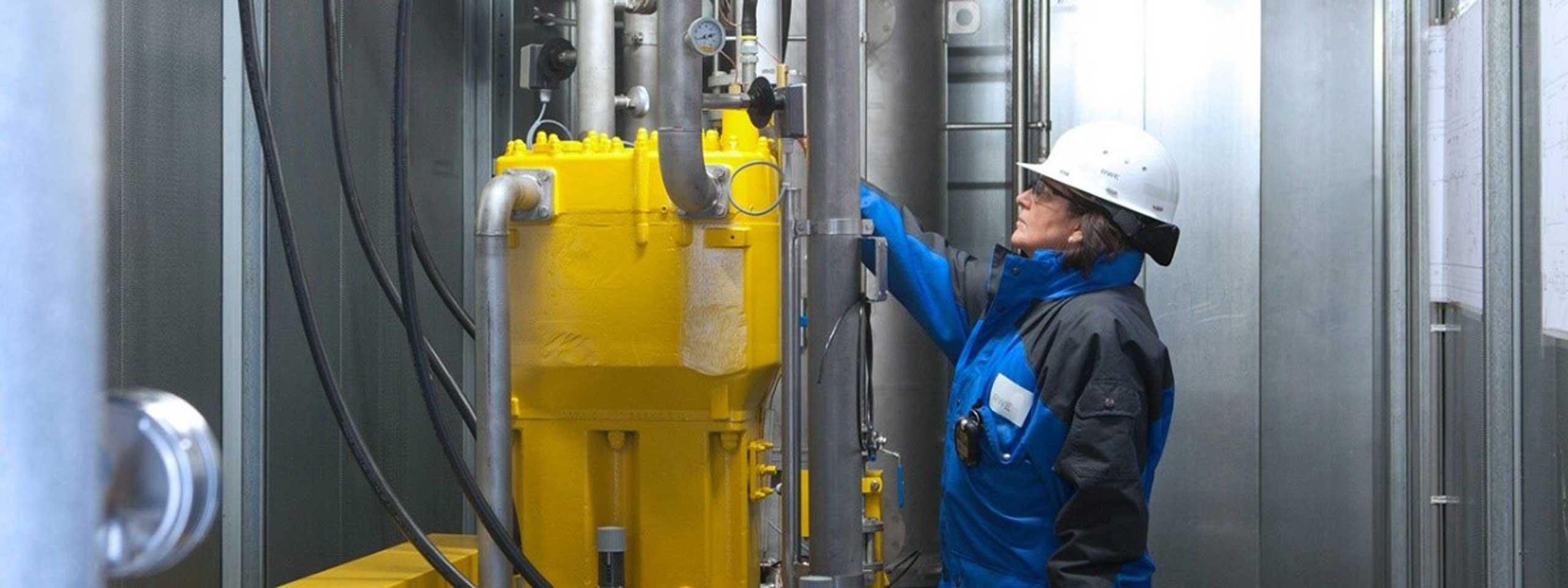 Eine Person in Schutzkleidung überprüft eine gelbe Maschine in einem industriellen Raum mit Metallwänden und Rohren.