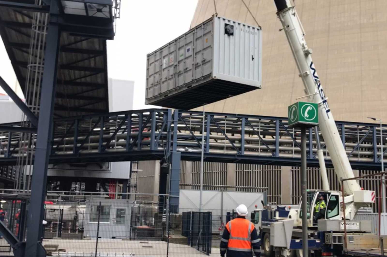 Ein Kran hebt einen Container an einer Baustelle über eine Metallkonstruktion. Arbeiter beobachten den Vorgang.