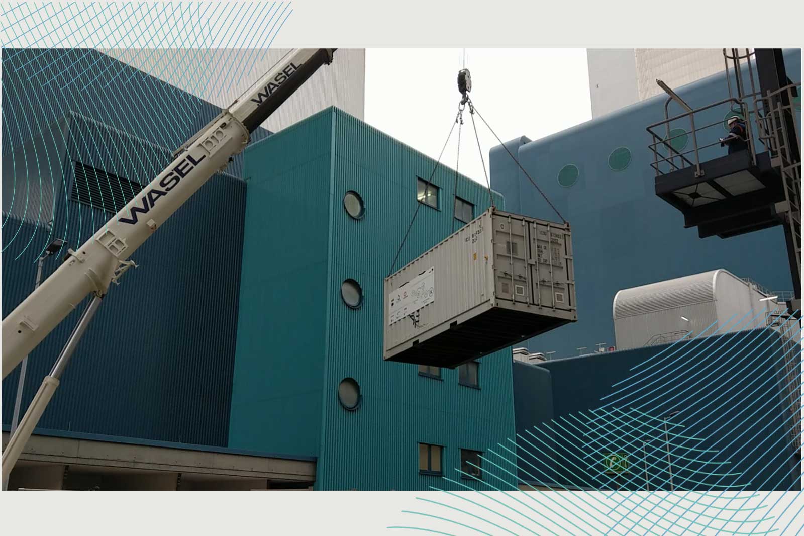 Ein Kran hebt einen Container über Gebäude mit blauen Fassaden und runden Fenstern, während ein Arbeiter auf einem Gerüst steht.