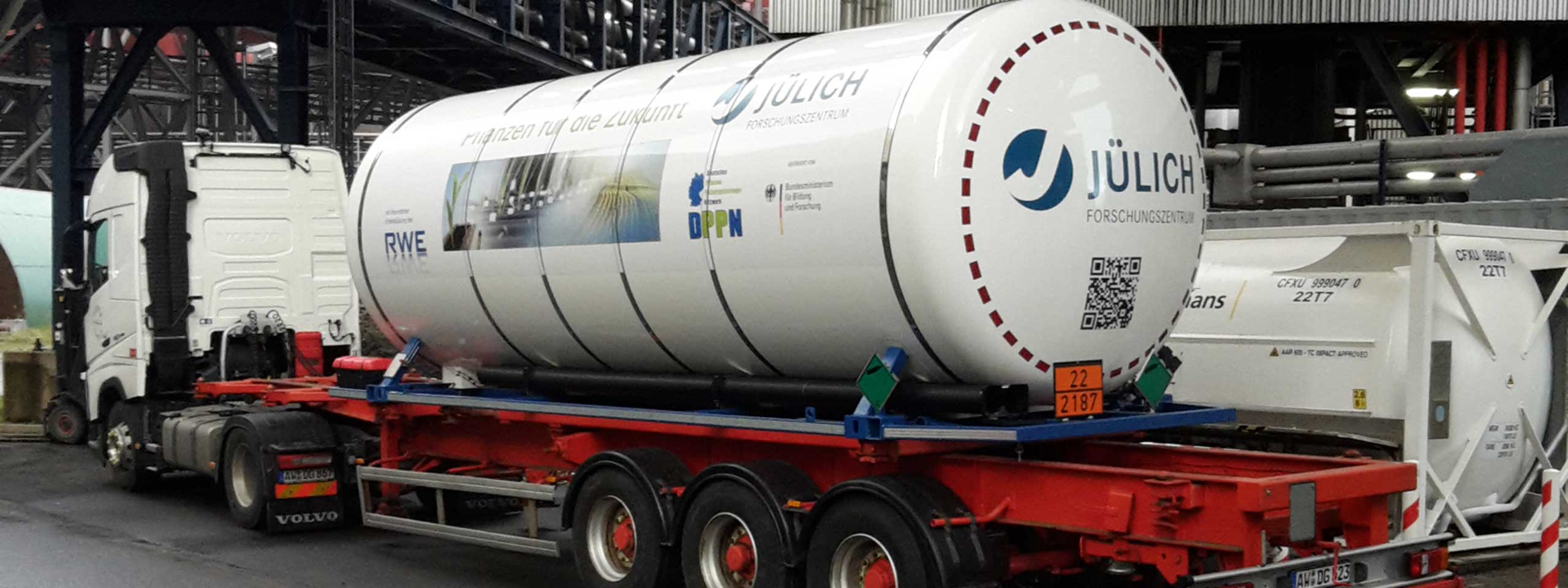 Ein Lastwagen transportiert einen zylindrischen Tank mit Logos und QR-Code, umgeben von Industrieanlagen.