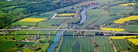 Eine Luftaufnahme einer Brücke über einen Fluss, umgeben von grünen Feldern und blühenden Rapsfeldern.