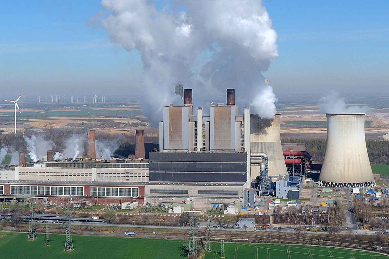 Ein Kraftwerk mit rauchenden Kühltürmen und Windrädern im Hintergrund. Grüne Felder dominieren die Landschaft.
