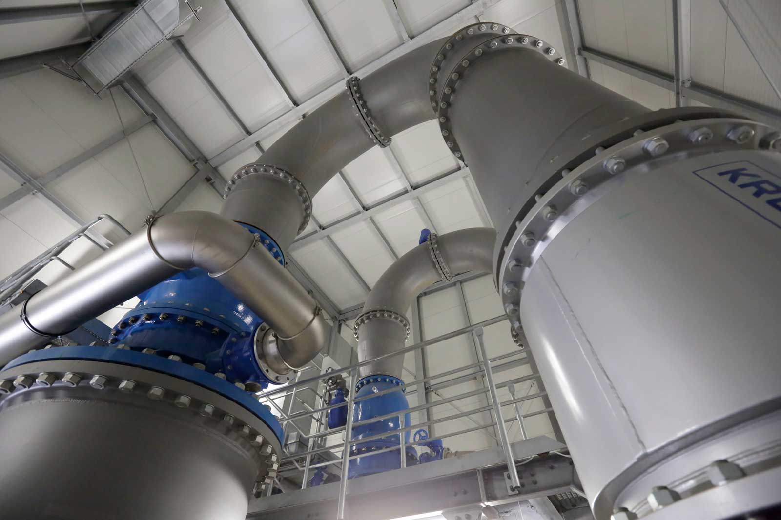 Ein Innenraum mit großen Metallrohren und einem blauen Ventil unter einem grauen Dach. Die Rohre sind ordentlich angeordnet.