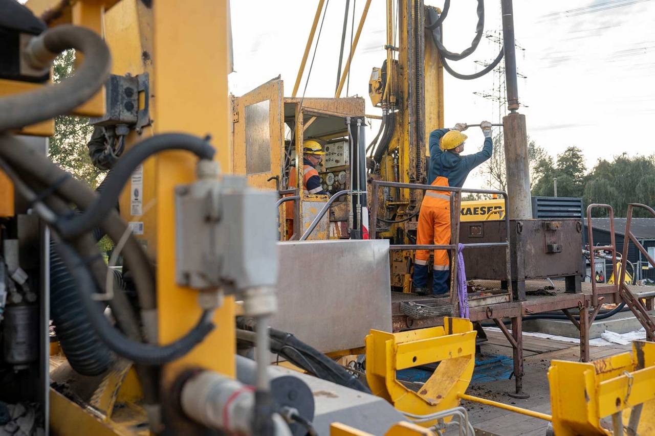 Zwei Arbeiter in Schutzkleidung bedienen eine Bohrmaschine auf einer Baustelle mit gelben Maschinen und einem Technikcontainer.