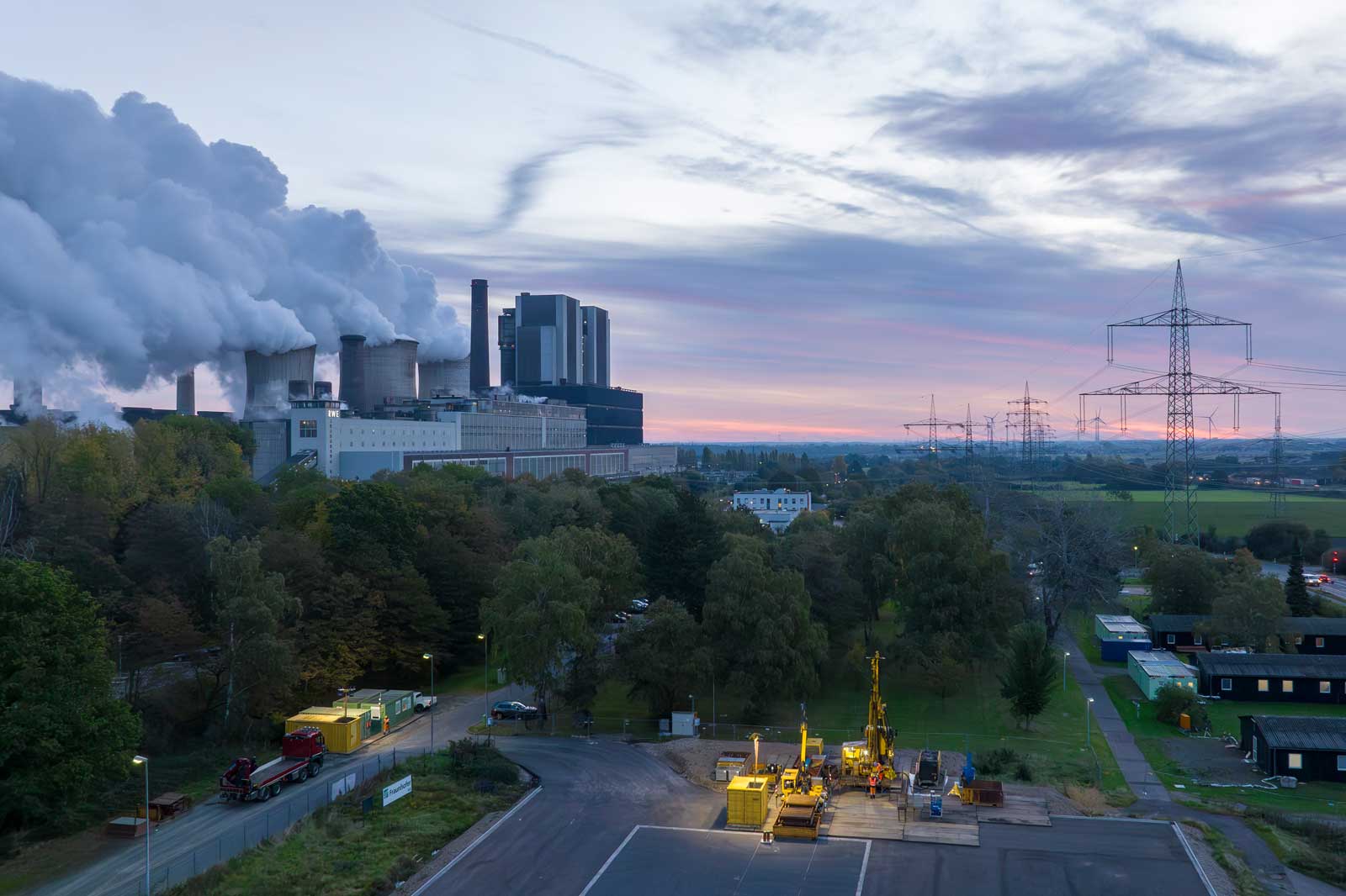 Ein Kraftwerk mit Rauchsäulen und Stromleitungen vor einem farbenfrohen Himmel. Betriebsstätten sind im Vordergrund.