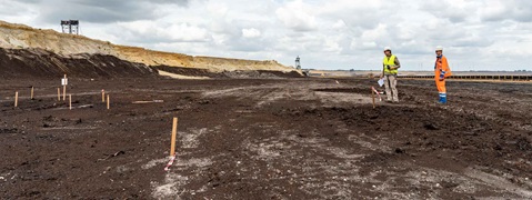 Zwei Arbeiter in Sicherheitswesten stehen auf einer braunen, unfruchtbaren Landschaft mit Markierungen, vor einer Kulisse aus Industriemaschinen.