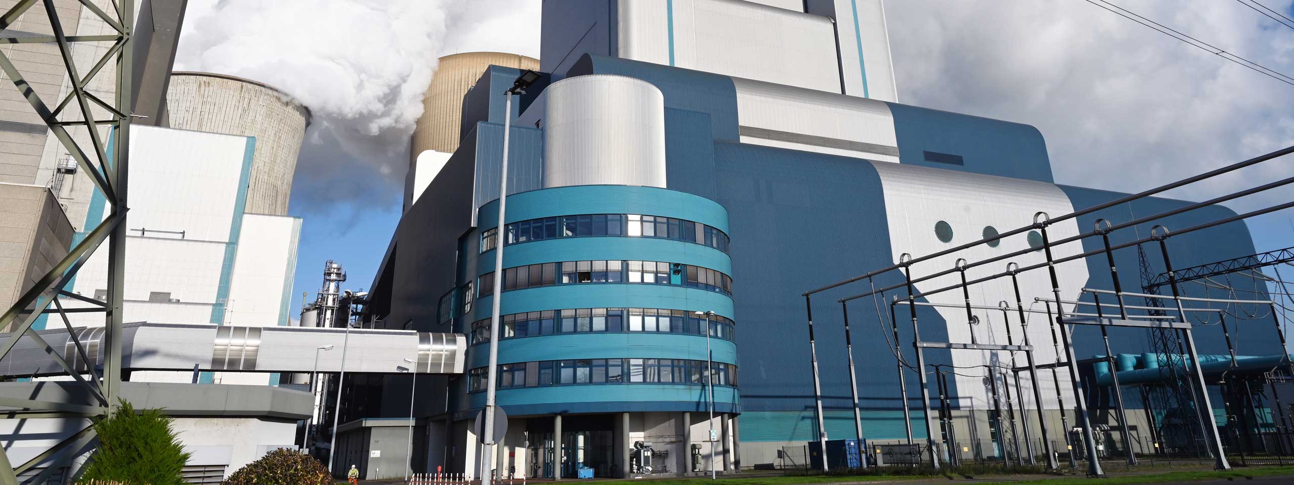 Ein modernes Kraftwerk mit großen Kühltürmen und einer blauen Fassade. Stromleitungen verlaufen im Vordergrund.