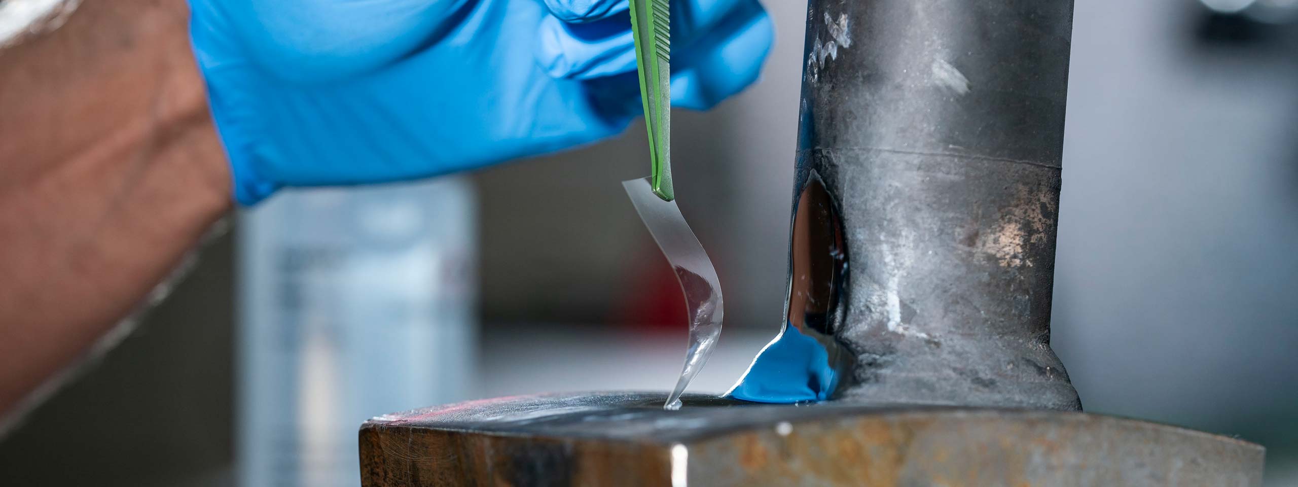 Eine behandschuhte Hand trägt mit einem Spatel einen klaren Kleber auf eine Metalloberfläche auf, im unscharfen Hintergrund.