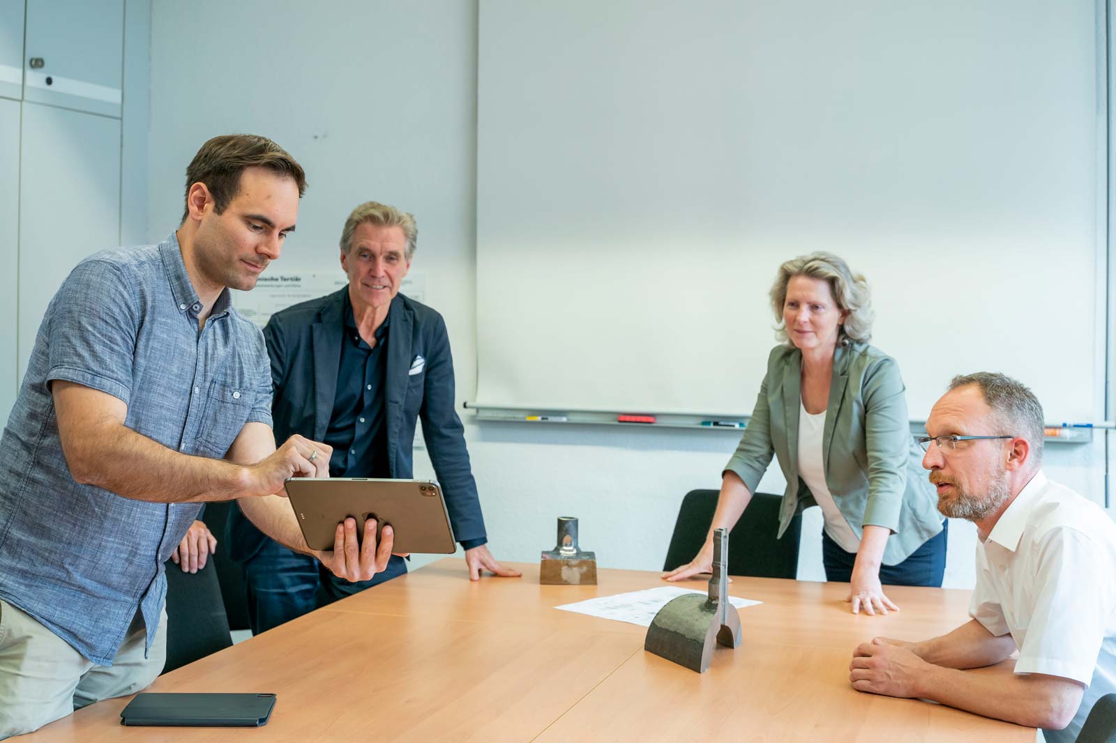 Eine Besprechung mit vier Personen am Tisch, während einer ein Tablet benutzt. Auf dem Tisch liegt ein Dokument.
