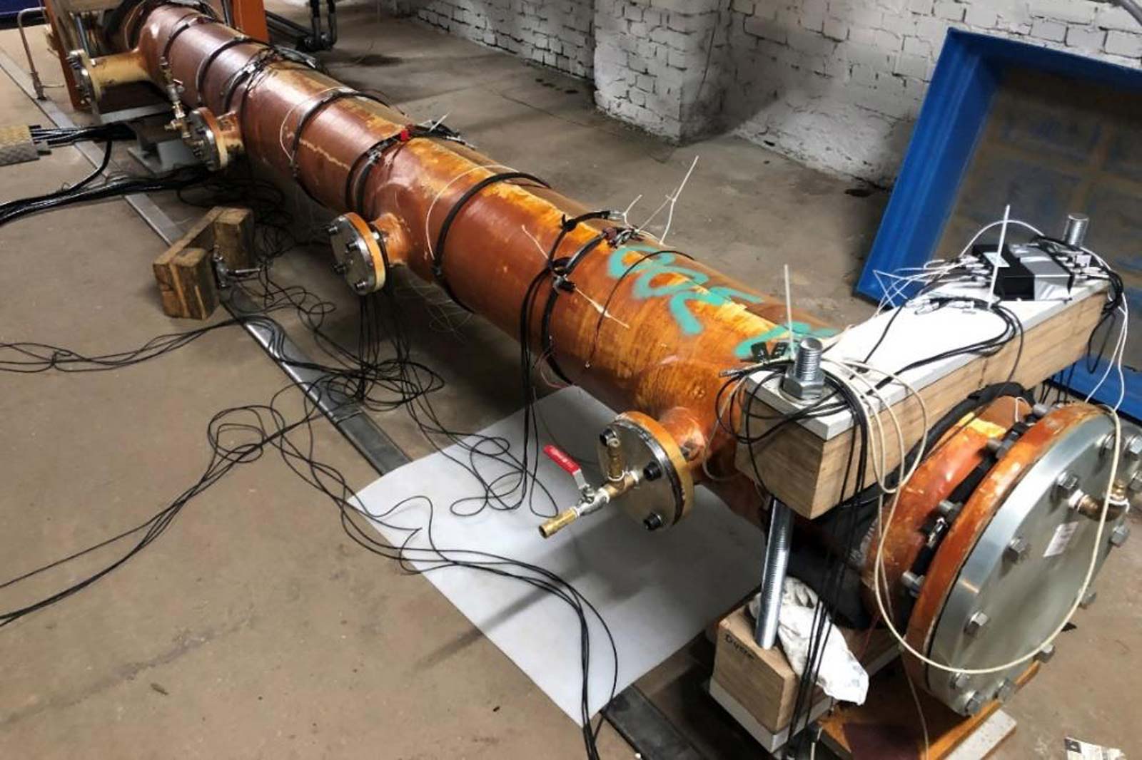 Ein großer, orangefarbener Zylinder mit vielen Kabeln und Ventilen, auf einem Betonboden in einem Lagerraum.